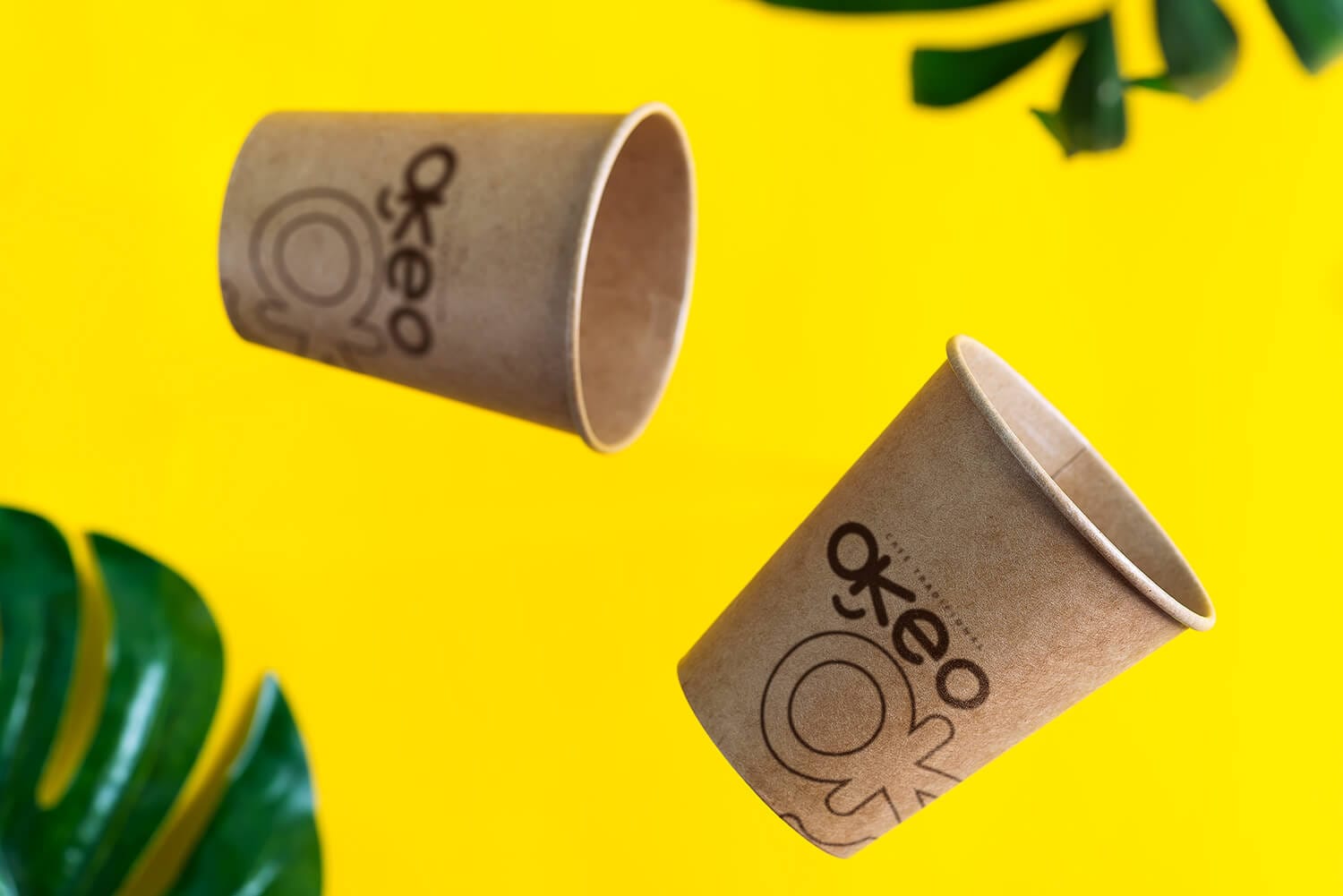 Dois copos de papel kraft com o logotipo da Okeo, em fundo amarelo com folhas verdes, representando design sustentável e identidade visual